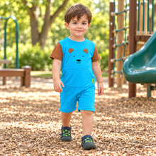 A toddler boy wearing a bright blue short-sleeve romper with brown sleeves and a bear face graphic, walking on a wood chip playground.