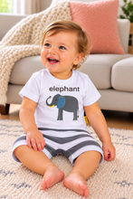 A smiling toddler sitting on a textured cream rug in a living room, wearing a white short-sleeve t-shirt with a gray elephant graphic and the word 'elephant' printed on the chest, paired with gray and white striped shorts.