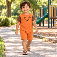 A young boy walking on a paved path in a sunny playground, wearing an orange short-sleeve romper with brown shoulder panels and a bear face graphic on the chest.