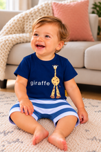 A smiling toddler sitting on a textured rug in a living room, wearing a navy blue t-shirt with a giraffe graphic and the word 'giraffe' printed on the front, paired with blue and white striped shorts.