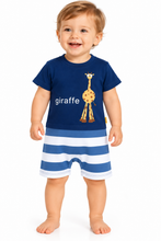 A smiling toddler stands against a white background wearing a navy blue short-sleeve top with a giraffe graphic and the word 'giraffe' in white, paired with blue and white horizontally striped shorts.