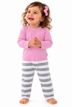 A toddler girl with curly brown hair wearing a pink long-sleeve top and gray and white striped pants, standing against a plain white background.