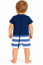 A baby viewed from the back wearing a short-sleeve romper with a solid navy blue top and blue and white striped shorts, standing against a white background.