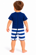 A baby viewed from the back wearing a navy blue short-sleeve romper with a vertical zipper and blue-and-white horizontally striped shorts, standing against a plain white background.